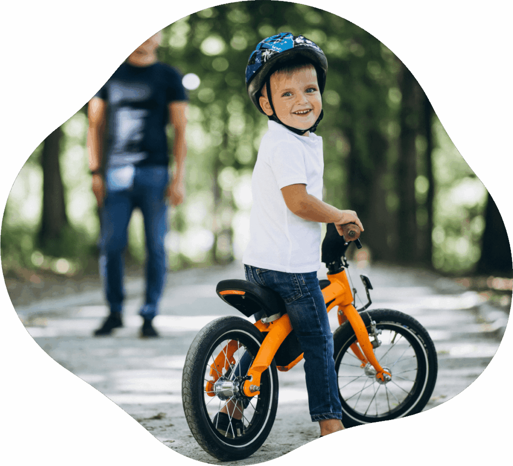 Junge in weißem Polohemd steht mit orangenem Kinderfahrrad im Park und lächelt